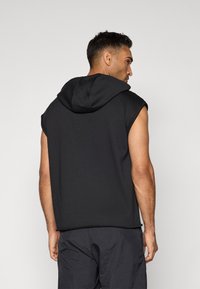 Svart ermeløs hettegenser med en stor hette, laget av mykt materiale. Har råkanter og en avslappet passform, brukt over mørke shorts.