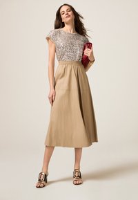 Beige midi rok met een elastische tailleband, gecombineerd met een glinsterende zilveren top met korte mouwen. Het model houdt een rode clutch vast en draagt zwarte sleehakken.