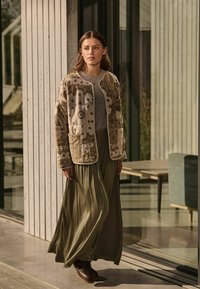 Beige geborduurd jack met bloemenpatronen, gecombineerd met een lange olijfgroene plissé rok en bruine veterschoenen. Houten achtergrond.