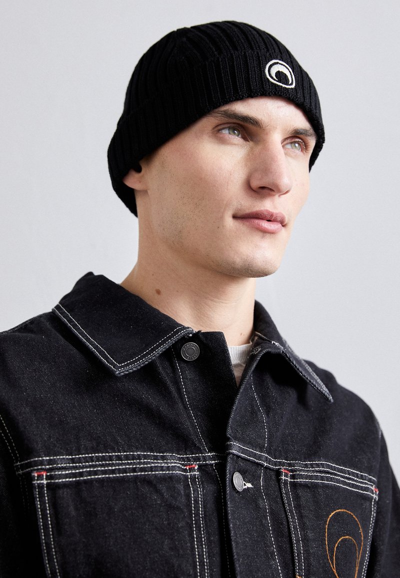 Căciulă neagră tricotată cu o textură ribbed și un logo circular. Pur și simplu cu o jachetă din denim neagră, prezentând cusături albe contrastante și nasturi argintii.