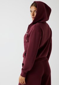 Vrouw in zijaanzicht die een bordeauxrode hoodie met de capuchon op en bijpassende broek draagt op een effen lichte achtergrond.