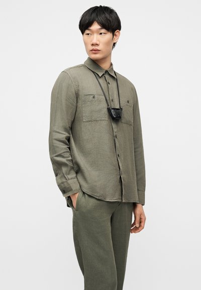 Νεαρός άνδρας που φοράει πουκάμισο με κουμπιά και παντελόνι σε ελιά πράσινο χρώμα, με μια μικρή μαύρη τσάντα να κρέμεται από κορδόνι γύρω από τον λαιμό του.