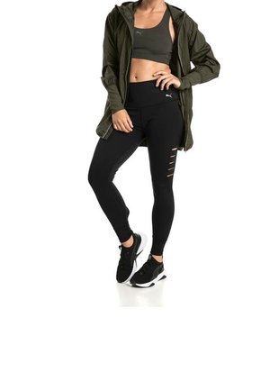 Schwarze Leggings mit Cut-Out-Details, grüner Sport-BH und grüner langer Jacke, ergänzt durch schwarze Sneakers mit weißen Akzenten.