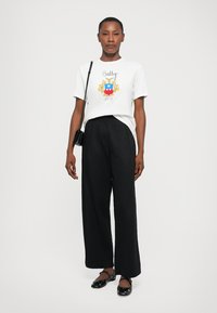 Woman standing wearing white graphic T-shirt, black wide-leg pants, black flats, and a small black crossbody bag, against plain background.