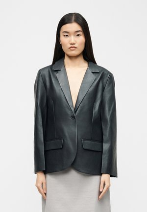 Žena s dlhými čiernymi vlasmi, oblečená v čiernom koženom blejzri na jedno zapínanie a svetlosivej sukni, stojaca na obyčajnom pozadí.