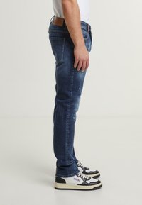 Mies, który ma na sobie wyblakłe niebieskie dżinsy i czarno-białe sneakersy, stoi bokiem na jednolitym jasnym tle.