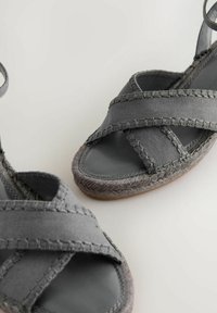 Šedé textilné sandále s prekríženými popruhmi a pletenou šnúrovou podrážkou. Majú hladkú stielku a prešité detaily pozdĺž okrajov.