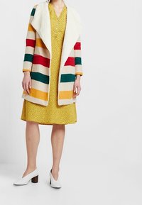 Donna che indossa un vestito giallo a fantasia, un cappotto a strisce multicolori con colletto in pile bianco e scarpe con tacco blocco bianche, in piedi su uno sfondo bianco.