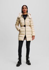 Beige pufferjas met een ceintuur in de taille, ritsluiting en capuchon. Voorzien van een gestikt ontwerp en zijzakken, gecombineerd met zwarte skinny jeans en laarzen.