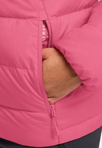 Rosa Puffjacke mit glatter Textur, ausgestattet mit einer seitlichen Reißverschlusstasche. Das Innenfutter zeigt eine weiche Fütterung. Akzente sind eine Kordel am Saum.