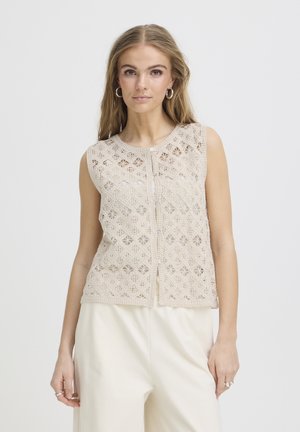 Frau mit langen blonden Haaren, die ein ärmelloses beiges Häkeloberteil mit Knöpfen über weißen Hosen trägt und vor einem einfarbigen Hintergrund steht.