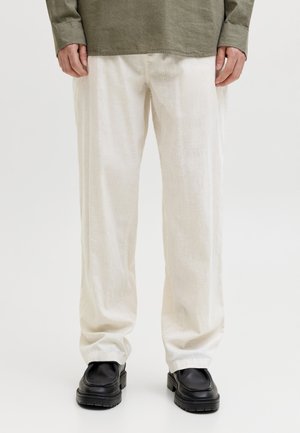 Άνδρας που φοράει άνετο λευκό παντελόνι από λινό, μαύρα χοντρά παπούτσια και μακρυμάνικη μπλούζα σε ελιά πράσινο χρώμα, στέκεται μπροστά σε απλό φόντο.