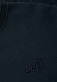 Sudadera verde oscura con cuello texturizado y acanalado. Presenta un logotipo bordado de "SuperDry" en púrpura oscuro en la esquina inferior izquierda.