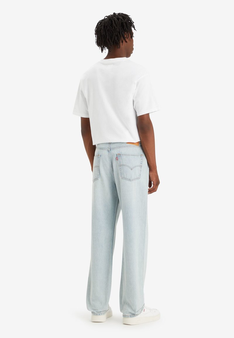 Camiseta blanca de manga corta combinada con jeans de corte holgado de color azul claro, con un parche de cuero en la parte trasera, y zapatillas blancas.