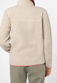 Beige fleecejacka med strukturerad tyg, hög krage och en rosa detalj i fållen. Har en söm över den övre ryggen.