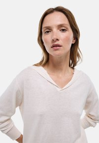 Femme aux cheveux châtain clair portant un pull crème à manches longues avec un col ample, debout les mains sur les hanches devant un fond uni.