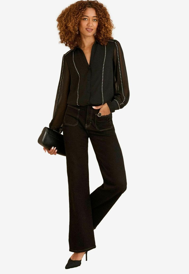 Blusa nera trasparente con chiusura a bottoni e accenti di borchie, abbinata a pantaloni neri a vita alta. Visibile una pochette nera e tacchi a punta.