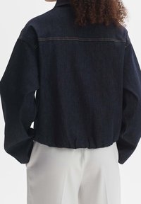 Mörk denimjacka med avslappnad passform, sänkt axelparti och elastisk midja. Har synlig sömnad och krage.