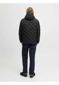Schwarze gepolsterte Jacke mit Kapuze und geometrischer Textur, kombiniert mit dunkelblauen Jeans. Ansicht von hinten, die das Design der Jacke betont.