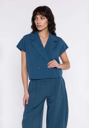 Kobieta o ciemnych włosach do ramion, ubrana w zielonkawy, krótki, dwurzędowy żakiet i dopasowane luźne spodnie, na białym tle.