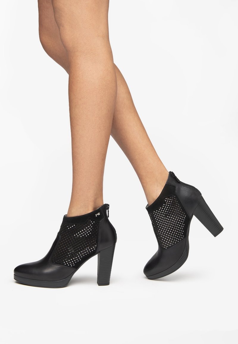 Nogi w czarnych botkach na wysokim obcasie, sięgających do kostki, z perforowanymi panelami z siateczki i bocznymi zamkami na białym tle.