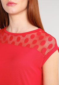 Červený top s pevným tělem a průhledným síťovým živůtkem s puntíkatým vzorem. Krátké rukávy a kulatý výstřih.