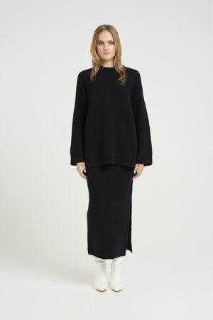 Black knitted sweater with long sleeves, paired with a black maxi skirt featuring a side slit. The model wears white ankle boots.