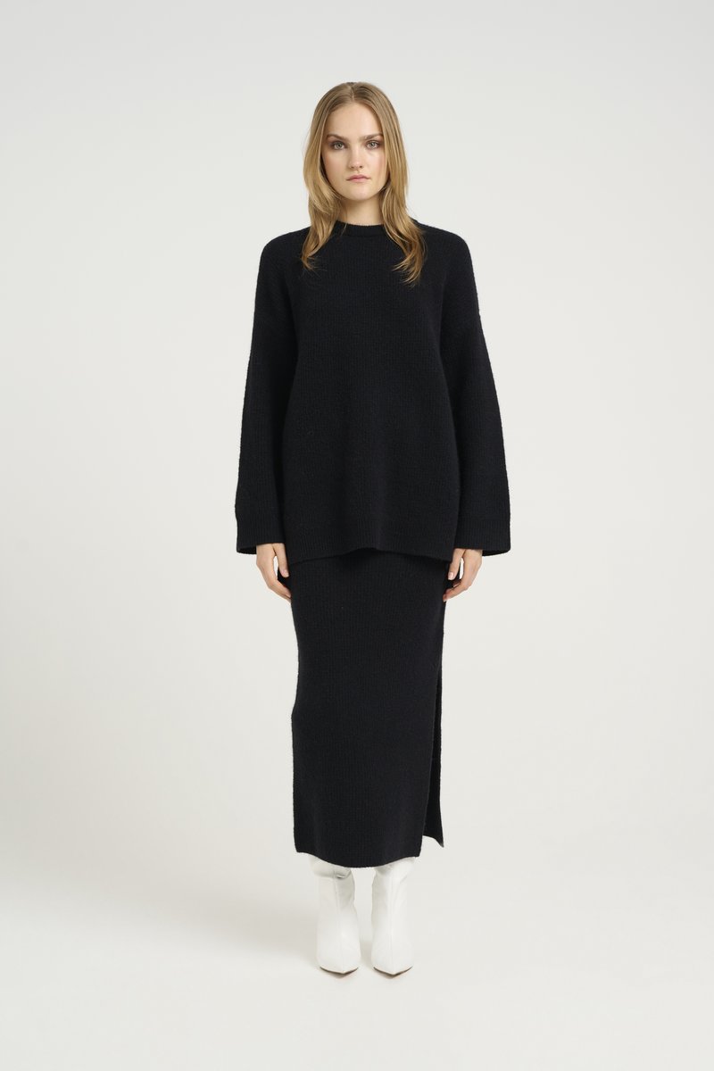 Pull en tricot noir à manches longues, associé à une jupe maxi noire avec une fente sur le côté. Le modèle porte des bottines blanches.