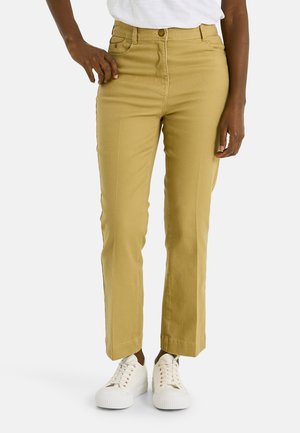 Persona vistiendo pantalones beige de pierna recta y zapatillas blancas, de pie con una mano apoyada cerca del bolsillo.