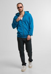 Niebieska bluza zapinana na zamek z kapturem na sznurku, w połączeniu z ciemnymi spodniami i beżowymi trampkami. Model ma okulary przeciwsłoneczne i pozuje z jedną ręką uniesioną.