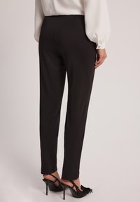 Czarne spodnie na miarę z prostą nogawką, wykonane z gładkiego materiału. Posiadają szew z tyłu i są zestawione z czarnymi sandałami na obcasie.