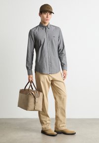 Camisa abotonada gris, pantalones beige, botas de ante marrón, sosteniendo un bolso marrón con detalles claros. Sombrero con el logo "BOSS" en la parte delantera.