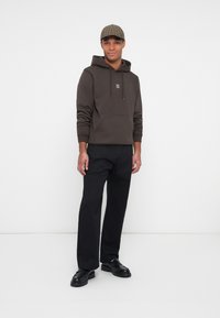 Hoodie marron avec une poche avant et capuche, affichant un petit logo. Associé à un pantalon noir et des chaussures noires. Casquette à motif écossais.