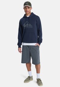 Sudadera azul marino con un gran logo gris, combinada con pantalones cortos grises. Una gorra verde y unos zapatos gris claros completan el conjunto.
