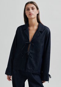 Jachetă din denim închis, cu mâneci lungi, guler cu colțuri și închideri frontale cu cravată. Țesătura are o textură netedă și o culoare albastră închisă.