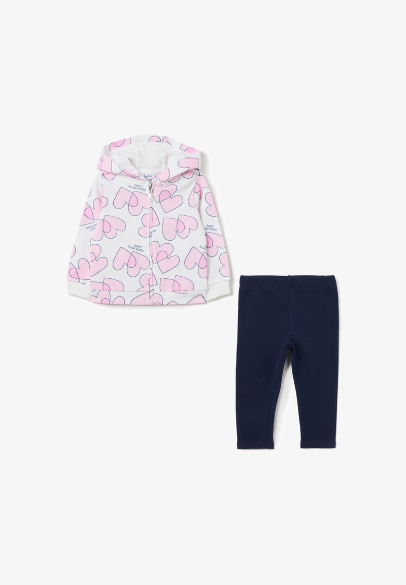 Giacca con cappuccio bianca con stampe a forma di cuore rosa, abbinata a leggings blu navy. La giacca presenta una chiusura con zip e polsini a coste.