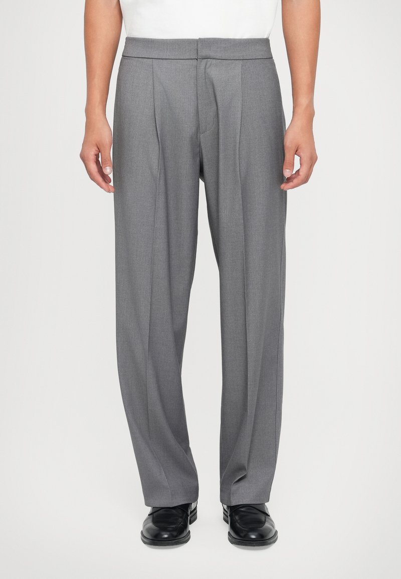 Pantalons tailleur gris avec une texture lisse, présentant des plis à l'avant et un design à jambe droite. Associés à des chaussures noires.