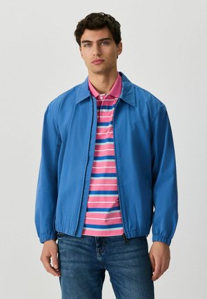 Junger Mann trägt eine blaue Jacke mit Reißverschluss über einem rosa-blau-weißen gestreiften Poloshirt und blauen Jeans, steht vor einem einfarbigen Hintergrund.