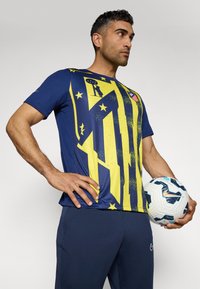 Mænds kortærmet blå trøje med gule grafikker, består af fede striber og stjerner. Bæres med mørke bukser og holder en mønstret fodbold.