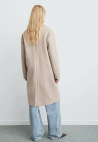 Manteau en laine beige avec une coupe droite, des manches longues et une couture centrale. Associé à un jean large bleu clair et porté avec des chaussures marron.