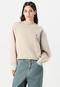 Felpa beige chiaro con maniche lunghe, scollo rotondo e piccolo logo. Abbinata a jeans verdi sbiaditi e dalle linee morbide.