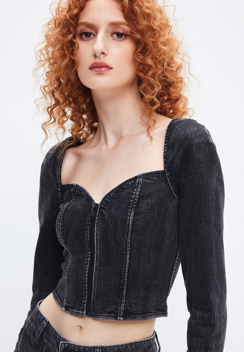Sort denim cropped jakke med firkantet udskæring, lange ærmer og lynlås foran. Har synlige sømme og en tætsiddende pasform.