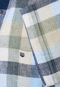 Tkanina w kratę w odcieniach niebieskiego, zielonego i beżowego. Posiada teksturowany splot oraz mały granatowy akcent z logo w pobliżu szwu.
