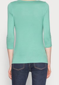 Hellgrünes, tailliertes Oberteil mit Dreiviertelärmeln, weicher Textur und weitem Ausschnitt; kombiniert mit dunkelblauen Jeans mit Rückensteppnähten.