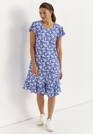 Kvinde i blå og hvid blomstret kjole med flæseærmer og kant, iført hvide sneakers, stående mod en ensfarvet hvid baggrund.