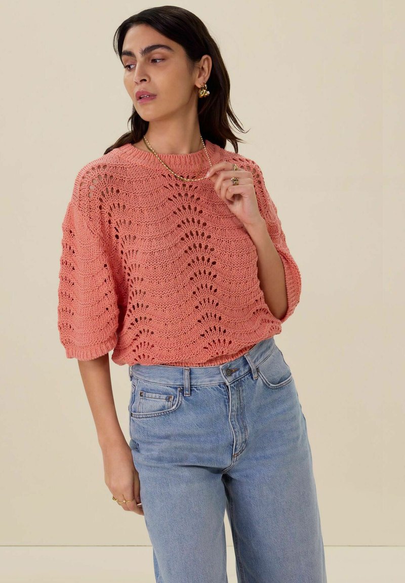 Koraalkleurige gebreide trui met een losse fit, korte mouwen en opengewerkte patroon, gecombineerd met lichtblauwe high-waisted jeans.