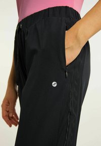 Czarne spodnie z wysokim elastycznym pasem, bocznymi kieszeniami i małym logo. Charakteryzują się gładką fakturą oraz kontrastowymi przeszyciami.