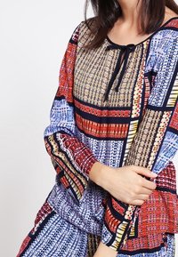 Blusa a fantasia multicolore con scollo regolabile, maniche a campana e intricati motivi geometrici nei toni del blu, rosso e oro.