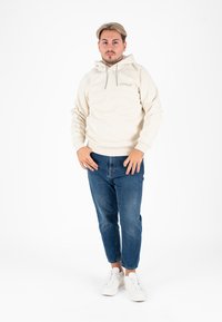 Beige hoodie met trekkoorden, geborduurd logo, gecombineerd met denim jeans en witte sneakers. Eenvoudige, casual outfit met een ontspannen pasvorm.