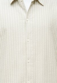 Κοντινό πλάνο μπεζ πουκαμίσου με υφή, κάθετες ρίγες και μυτερό γιακά πάνω από ένα λευκό κοντομάνικο.